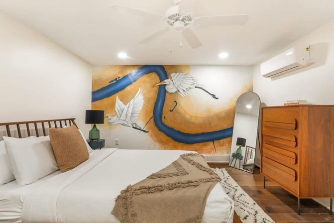 Modern bedroom interior with white bed and tan throw, wooden headboard, blue river and flying heron wall mural, full-length mirror and mid-century wooden dresser