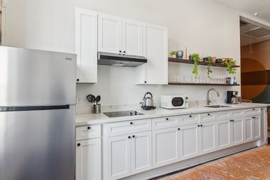 Bright modern white kitchen with stainless refrigerator, marble-look countertop, built-in electric cooktop, kettle and microwave, open wood shelves with glasses and trailing plants, sink and coffee maker