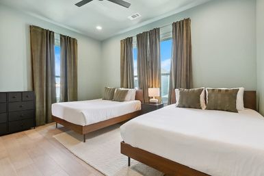 Modern guest bedroom with two wooden platform beds and white linens, olive curtains, bedside lamp, black dresser and ceiling fan
