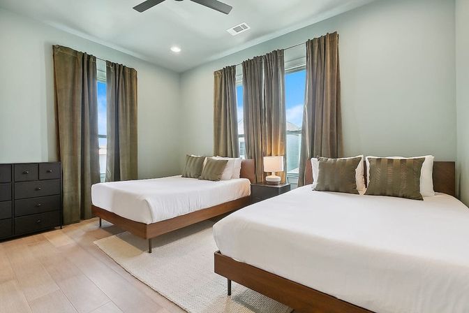 Modern guest bedroom with two wooden platform beds and white linens, olive curtains, bedside lamp, black dresser and ceiling fan