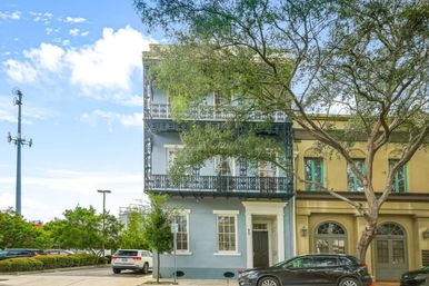 Three-story historic rowhouse with ornate wrought-iron balconies and pale blue facade on a tree-lined urban street, parked cars and bright sky in the background.