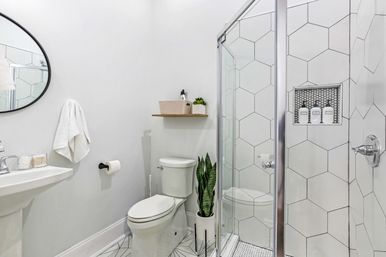 Bright modern white bathroom with glass-enclosed hexagon-tile shower, pedestal sink, round mirror, toilet, wooden shelf and potted snake plant