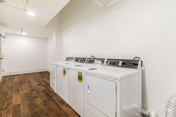 Bright communal laundry room with a row of white commercial washers and dryers along a white wall, dark wood-look flooring, exposed ceiling pipes and overhead lighting.