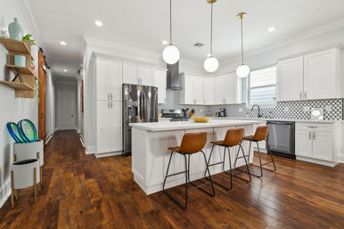 Bright modern white kitchen with large island and three leather bar stools, stainless steel appliances, hexagon tile backsplash, globe pendant lights and warm hardwood floors.