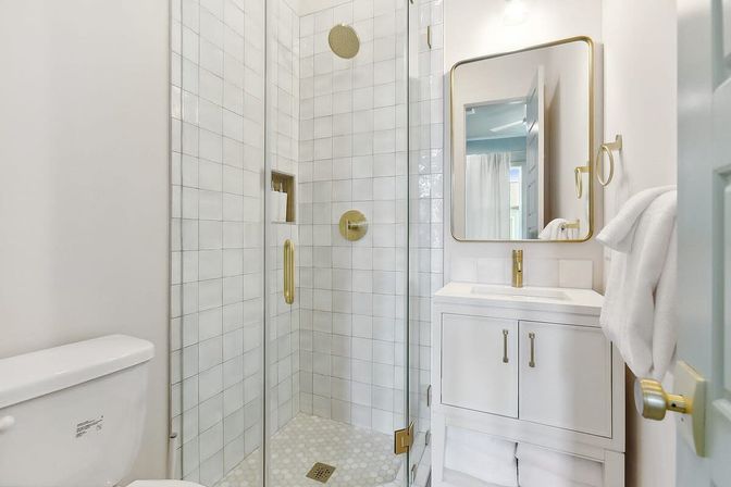 Bright, spa-like white bathroom with glass-enclosed tiled shower, brass fixtures, white vanity and folded towels