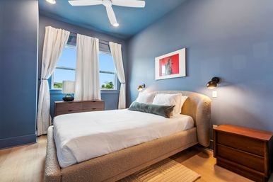 Bright modern blue bedroom with upholstered bed, white linens and green bolster, wooden nightstands, wall sconces, ceiling fan, and sunlit window with white curtains — cozy coastal-style retreat.