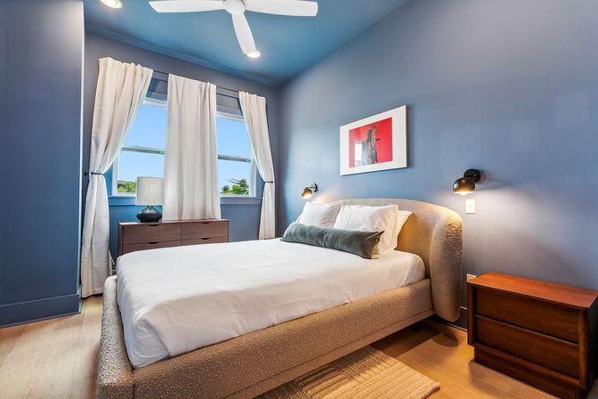Bright modern blue bedroom with upholstered bed, white linens and green bolster, wooden nightstands, wall sconces, ceiling fan, and sunlit window with white curtains — cozy coastal-style retreat.