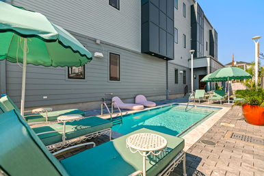 Sunny outdoor swimming pool in a modern urban courtyard with teal lounge chairs, scalloped green umbrellas, round white side tables, and potted plants beside a gray multi-story building.
