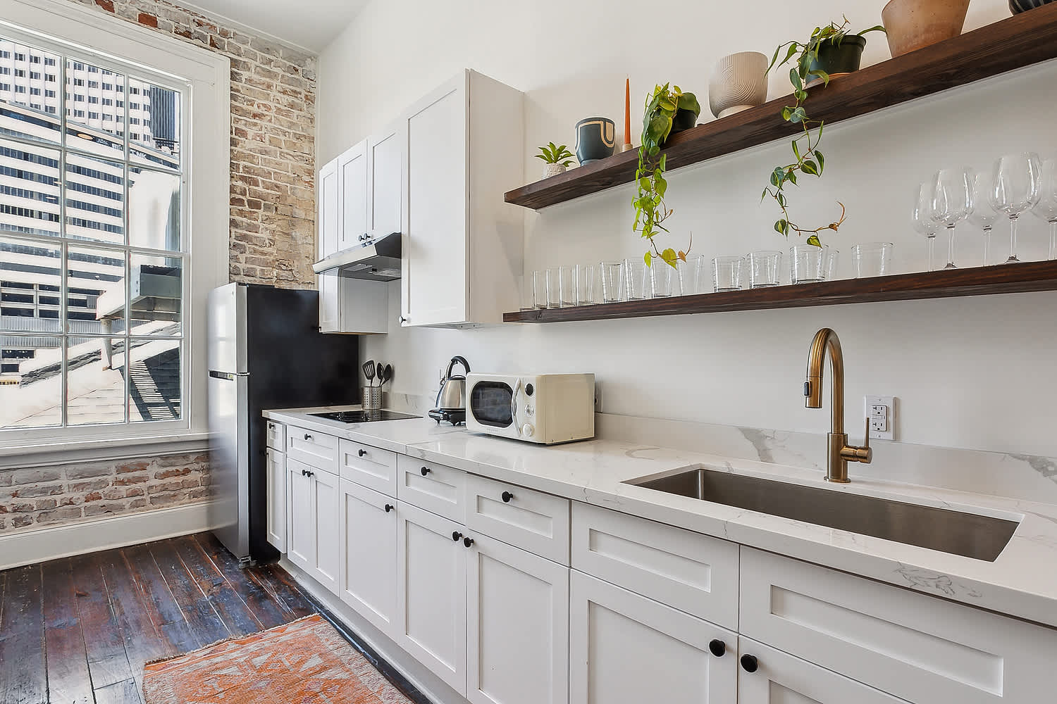 Bright urban loft kitchen with exposed brick and city-view window, white shaker cabinets, marble countertop, brass faucet, open wooden shelves with plants and glassware, stainless fridge and dark hardwood floors.