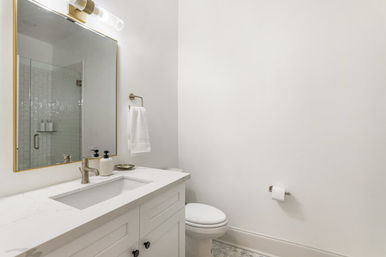 Bright, spa-like modern white bathroom with quartz vanity and undermount sink, gold-framed mirror and tubular sconce, towel ring, toilet, and glass-enclosed subway-tile shower — minimalist contemporary design.
