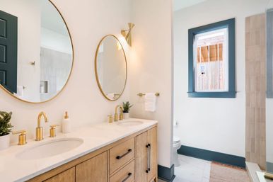 Bright modern bathroom with double vanity, wood cabinets, white quartz countertop, gold faucets and round mirrors, blue-trim window and walk-in shower