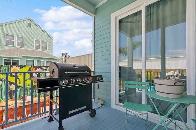 Sunny coastal balcony deck with black gas grill, mint-green bistro table and chairs with a bucket of chilled drinks, sliding glass doors, and a colorful floral fence mural