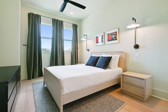 Cozy sunlit modern bedroom with light wood bed and nightstand, crisp white bedding and navy accent pillows, dark green curtains over twin windows, black ceiling fan, wall sconces and colorful framed art.