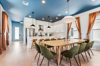 Open-plan modern kitchen and dining room with a blue ceiling, light wood floors, long wooden table surrounded by olive-green chairs, white island with wooden bar stools, stainless steel appliances, pendant lights, and rust-colored curtains.
