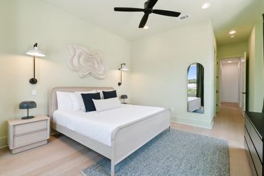 Sunlit modern master bedroom with pale green walls, king bed in white linens and navy accent pillows, light wood nightstands with lamps, wall sconces, black ceiling fan, arched full-length mirror and gray area rug.