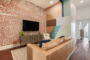 Urban loft-style living room with tan leather sofa, exposed red brick wall and wall-mounted TV, mid-century console, colorful geometric mural, patterned rug, hardwood floors and doorway to a balcony.