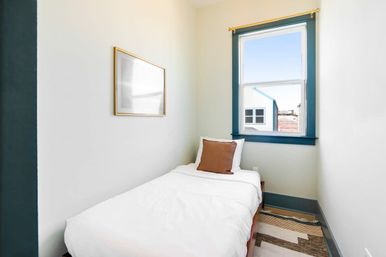 Cozy compact single bedroom with white bedding and brown accent pillow, teal-trim window letting in daylight and partial urban view, simple framed art and patterned rug.