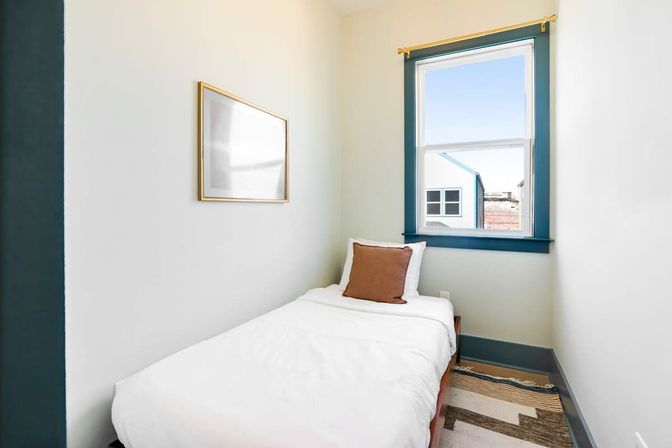 Cozy compact single bedroom with white bedding and brown accent pillow, teal-trim window letting in daylight and partial urban view, simple framed art and patterned rug.