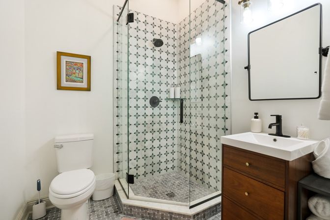 Bright modern bathroom interior with corner glass shower featuring geometric patterned tiles and black fixtures, mosaic floor, white toilet, and wooden vanity with black faucet and square mirror.