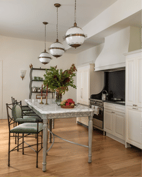 Bright neutral kitchen with marble island, carved apron and green cushioned metal bar stools, three vintage globe pendant lights, white cabinets, stainless range with black backsplash and hardwood floors