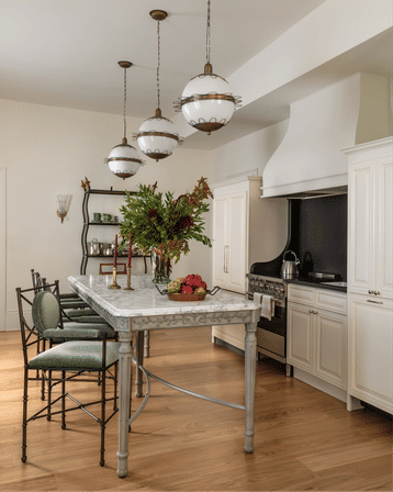 Bright neutral kitchen with marble island, carved apron and green cushioned metal bar stools, three vintage globe pendant lights, white cabinets, stainless range with black backsplash and hardwood floors