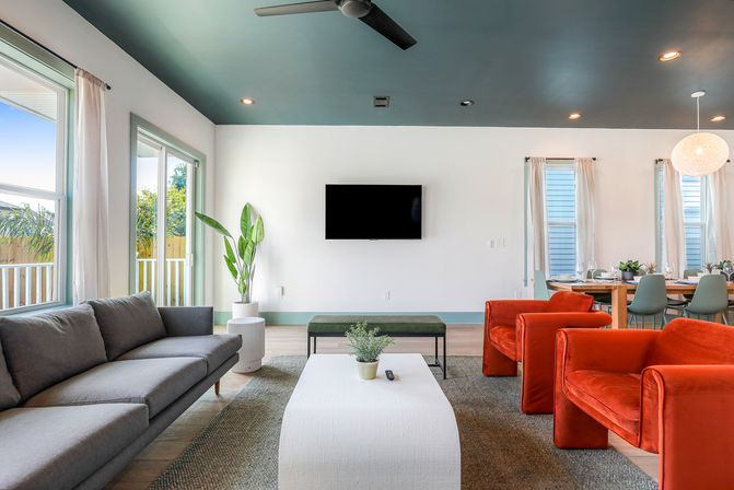 Modern open-plan living room with teal ceiling, gray sofa and two vibrant orange armchairs facing a wall-mounted TV, white coffee table, potted plant, and a bright dining area with large windows.