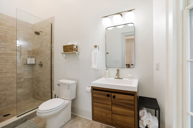 Bright modern home bathroom with glass walk-in shower and beige subway tile, white toilet, walnut wood vanity with brass faucet, rectangular mirror and globe vanity lights, wicker shelf and rolled towels