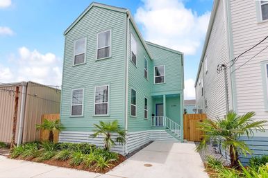 Cheerful mint-green two-story coastal-style house with vinyl siding, front steps and small porch, concrete driveway and tropical landscaping with palm plants under a bright blue sky.