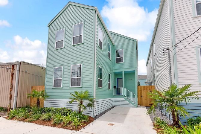 Cheerful mint-green two-story coastal-style house with vinyl siding, front steps and small porch, concrete driveway and tropical landscaping with palm plants under a bright blue sky.