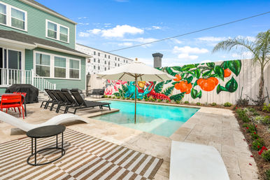 Sunny backyard pool and patio at a mint-green house, featuring a white umbrella over a shallow ledge, black lounge chairs, red patio chairs, striped outdoor rug, and a vibrant tropical mural of oranges and flowers on the fence.