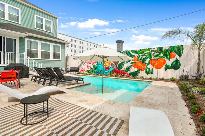 Sunny backyard pool and patio at a mint-green house, featuring a white umbrella over a shallow ledge, black lounge chairs, red patio chairs, striped outdoor rug, and a vibrant tropical mural of oranges and flowers on the fence.