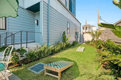 Sunny narrow side yard beside a blue-gray house with tropical plants, patio seating and two cornhole boards on a green lawn