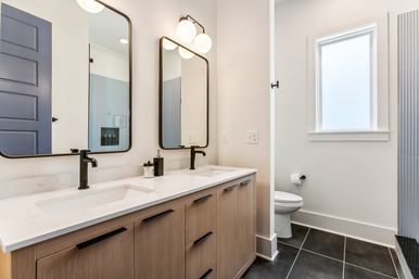 Bright modern bathroom with double quartz sink vanity, light wood cabinets, matte-black faucets and hardware, rounded rectangular mirrors with globe lights, frosted window, toilet alcove and dark floor tiles.
