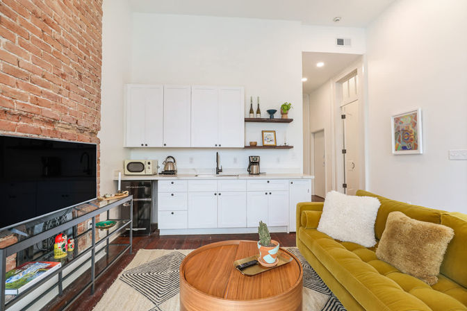 Bright loft-style living room with white kitchenette, exposed brick wall, mustard-green velvet sofa, round wooden coffee table with potted plant, flat-screen TV and hardwood floors