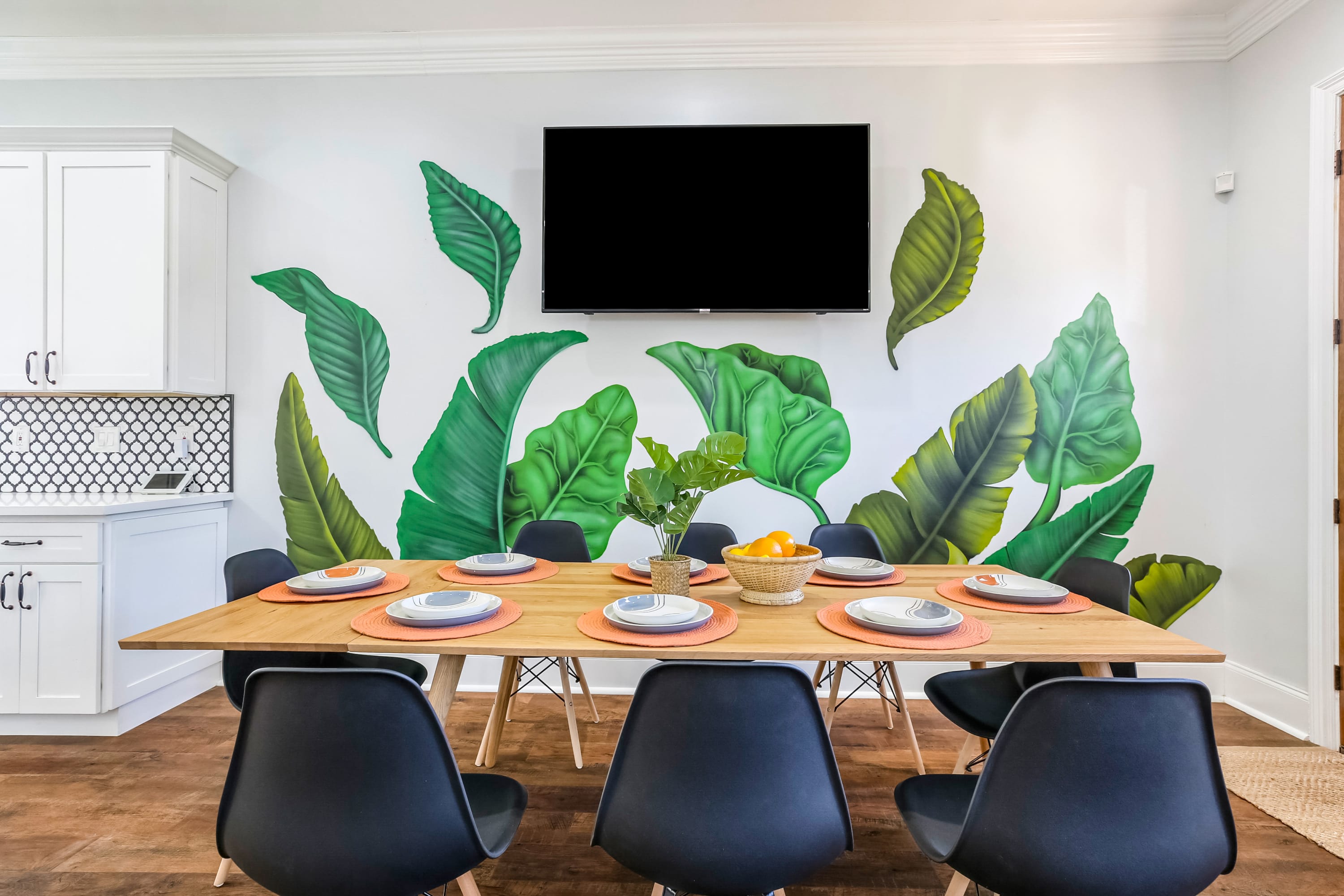 Modern dining area with wooden table set for six, black chairs, vibrant tropical leaf wall mural, wall-mounted TV, plant centerpiece and white kitchen cabinets in an open-concept interior