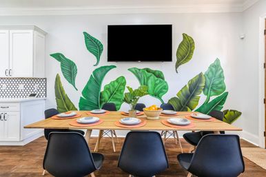 Modern dining area with wooden table set for six, black chairs, vibrant tropical leaf wall mural, wall-mounted TV, plant centerpiece and white kitchen cabinets in an open-concept interior