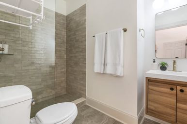 Bright modern bathroom with glass walk-in shower and gray subway tile, white toilet, wood vanity with brass faucet and mirror, neatly folded white towels on a wall rack and a small potted plant.