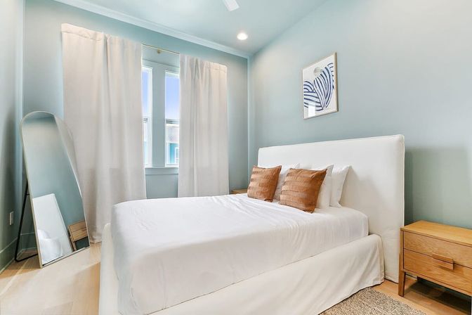 Light-filled modern bedroom with coastal blue walls, white upholstered bed, two tan throw pillows, wooden nightstand and full-length leaning mirror.