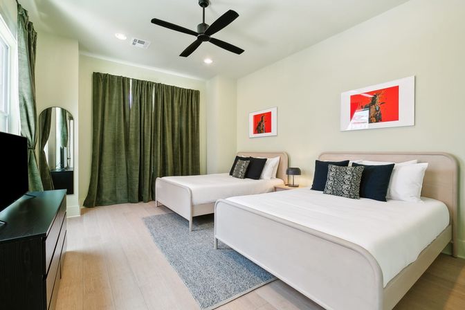 Contemporary twin bedroom in an urban rental with two light-wood beds, crisp white linens, black accent pillows, dark green floor-to-ceiling curtains, black ceiling fan, red-accented wall art, TV on a black dresser and a gray area rug.