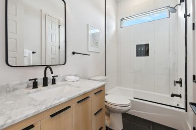 Bright modern bathroom with marble countertop, oak vanity, matte black fixtures and glass-enclosed tub-shower with high horizontal window
