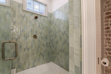 Walk-in bathroom shower with glossy seafoam-green subway tiles, small window, mosaic white floor, brushed-nickel showerhead and glass door handle, and an exposed brick accent wall.