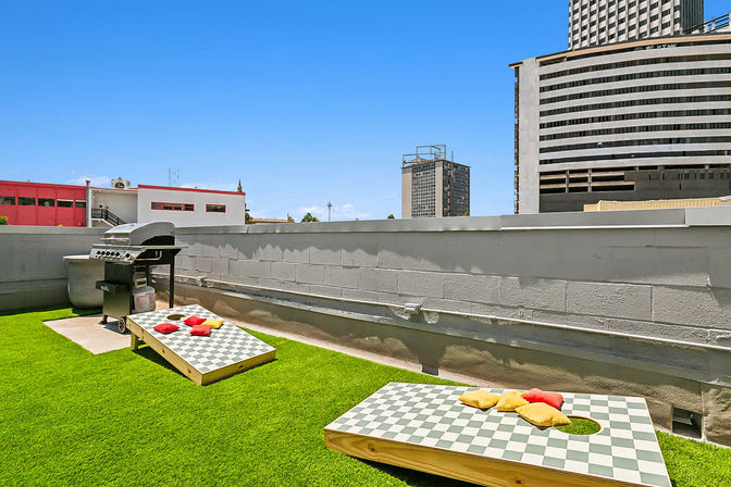 Urban rooftop patio with bright green turf, two checkered cornhole boards with colorful beanbags, a stainless-steel grill, concrete parapet and downtown high-rise skyline under a clear blue sky.