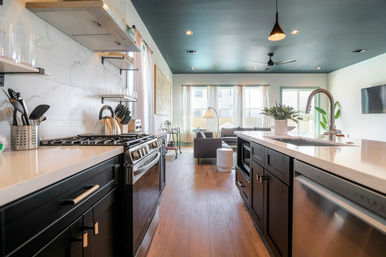 Bright modern open-concept kitchen and living area in a suburban home — dark cabinets, quartz island with sink, stainless gas range and dishwasher, teal ceiling with pendant light, wood floors, and a sunlit sofa by large windows.