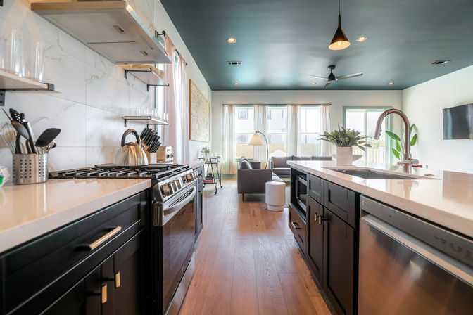 Bright modern open-concept kitchen and living area in a suburban home — dark cabinets, quartz island with sink, stainless gas range and dishwasher, teal ceiling with pendant light, wood floors, and a sunlit sofa by large windows.