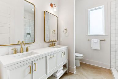 Bright modern white bathroom featuring a double-sink vanity with gold faucets and mirrors, a white countertop, towel rack, toilet, and tiled shower illuminated by a window.