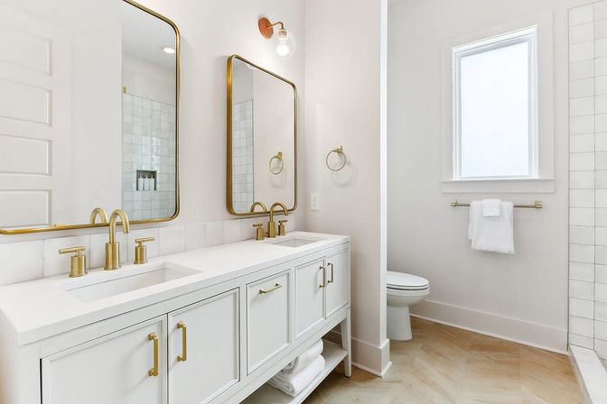 Bright modern white bathroom featuring a double-sink vanity with gold faucets and mirrors, a white countertop, towel rack, toilet, and tiled shower illuminated by a window.