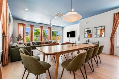 Sunlit modern open-plan dining and living room with a long wooden table surrounded by olive-green chairs, sectional sofa by large windows and sliding glass door showing a backyard, blue painted ceiling, pendant light, hardwood floors and colorful wall art.
