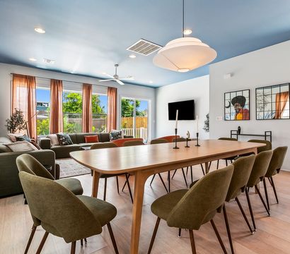 Sunlit modern open-plan dining and living room with a long wooden table surrounded by olive-green chairs, sectional sofa by large windows and sliding glass door showing a backyard, blue painted ceiling, pendant light, hardwood floors and colorful wall art.