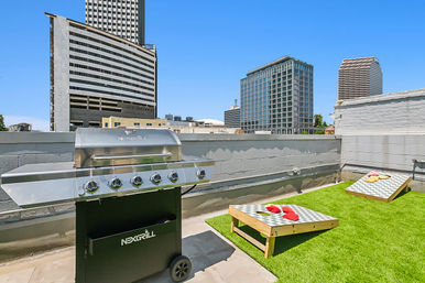 Sunny downtown rooftop patio with stainless-steel gas grill, artificial turf, two cornhole boards with colorful bean bags, and surrounding high-rise buildings