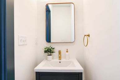 Small modern powder room with white sink on a dark vanity, gold faucet and towel ring, rounded-rectangle mirror, small potted plant on the counter, and bright white walls.